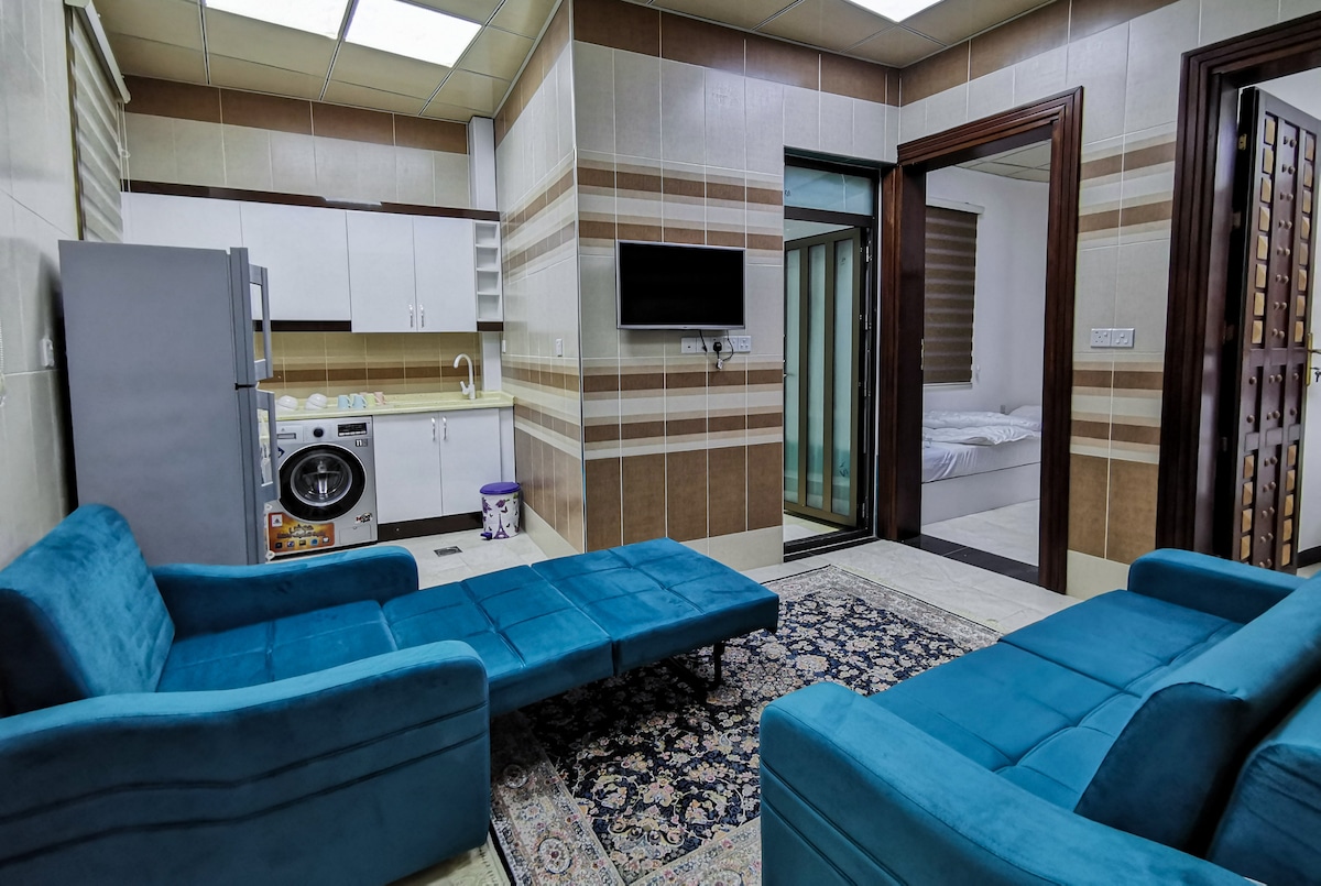 A modern living area features two plush sofas in shades of blue facing each other. A small area rug is placed on the tile floor. The kitchen area includes a refrigerator, washer, and a wall-mounted TV. Natural light enters through sliding glass doors leading to the bedrooms.