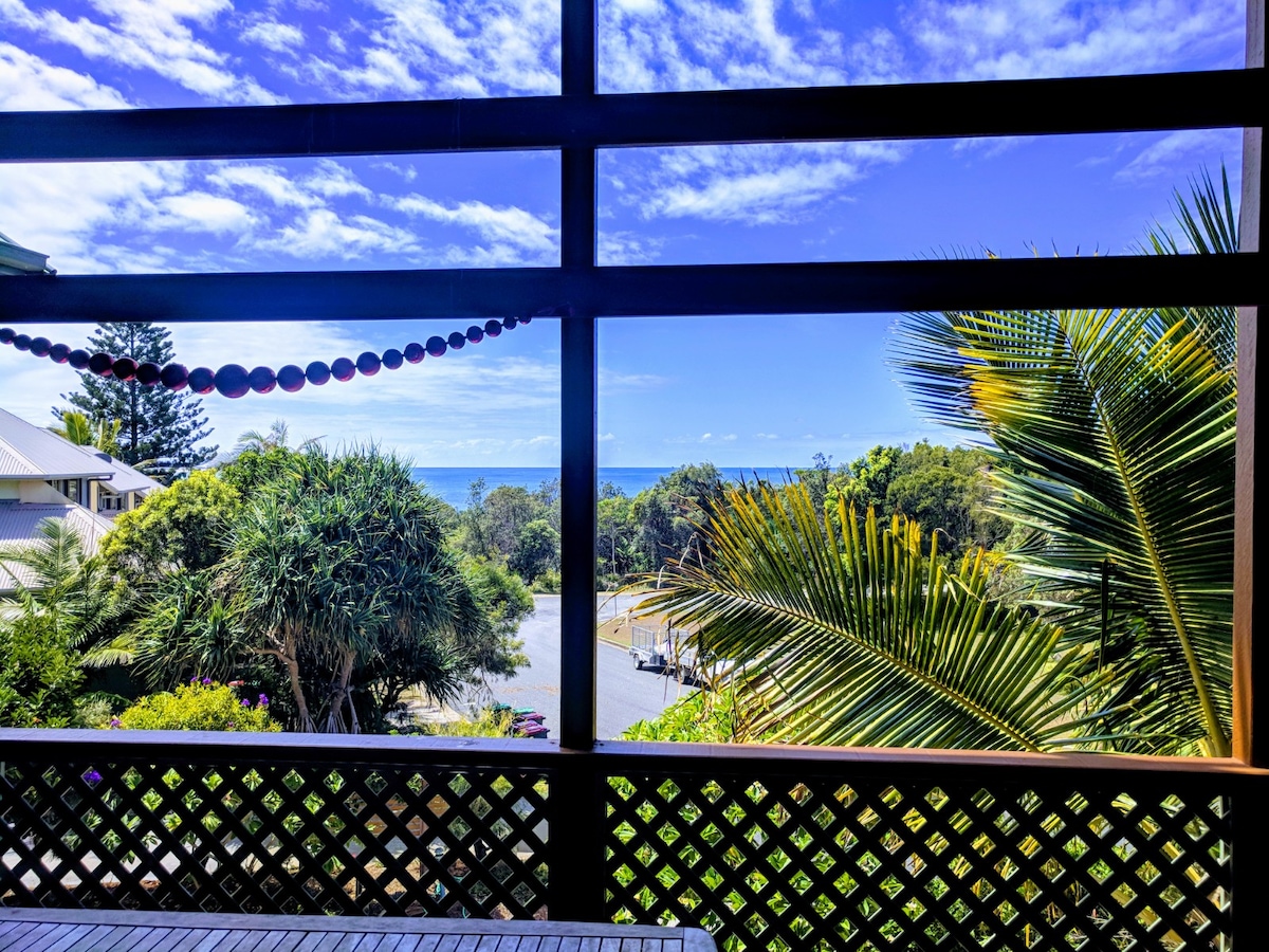 An ocean view is framed by large glass panels, offering a glimpse of the vibrant landscape. Lush greenery and palm fronds can be seen in the foreground, while the sparkling blue water of Spooky Beach is visible in the distance under a bright sky.