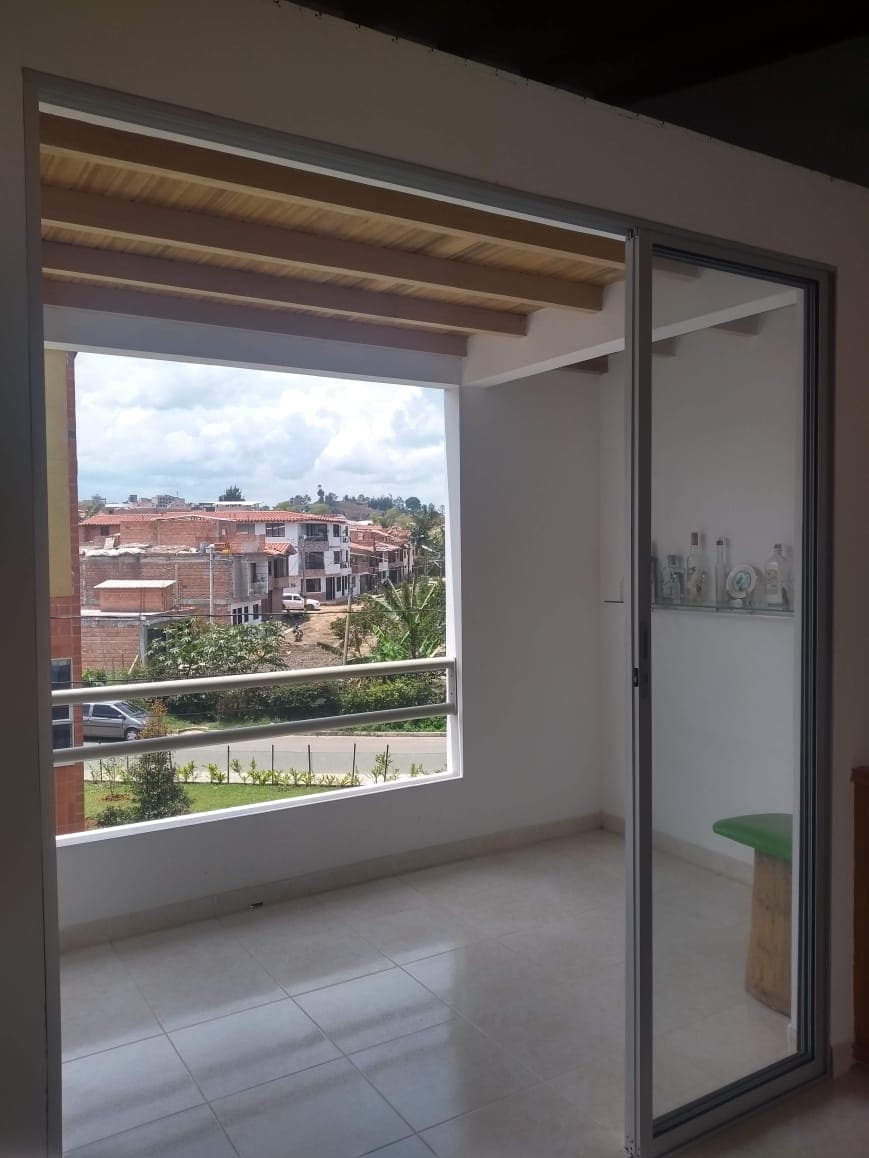 A bright balcony area is enclosed by sliding glass doors, offering views of urban rooftops and greenery. The space features a small wooden stool and a minimalist aesthetic, with smooth tile flooring and natural light streaming in from the open door.