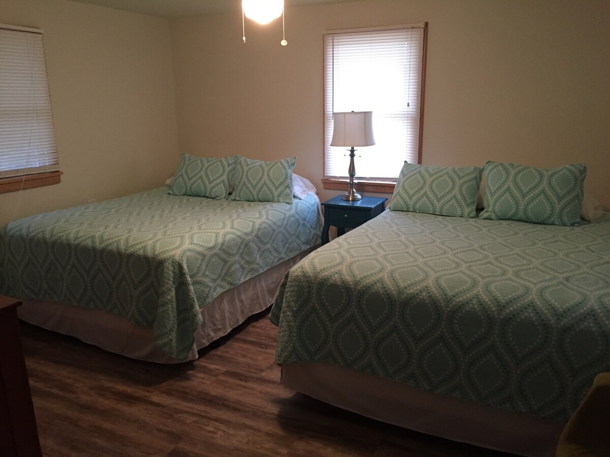 A room featuring two queen-sized beds, each dressed in light teal patterned bedding. A bedside lamp sits atop a small blue nightstand, providing soft illumination. Natural light filters in through two windows, accompanied by light beige walls and a warm wood floor.
