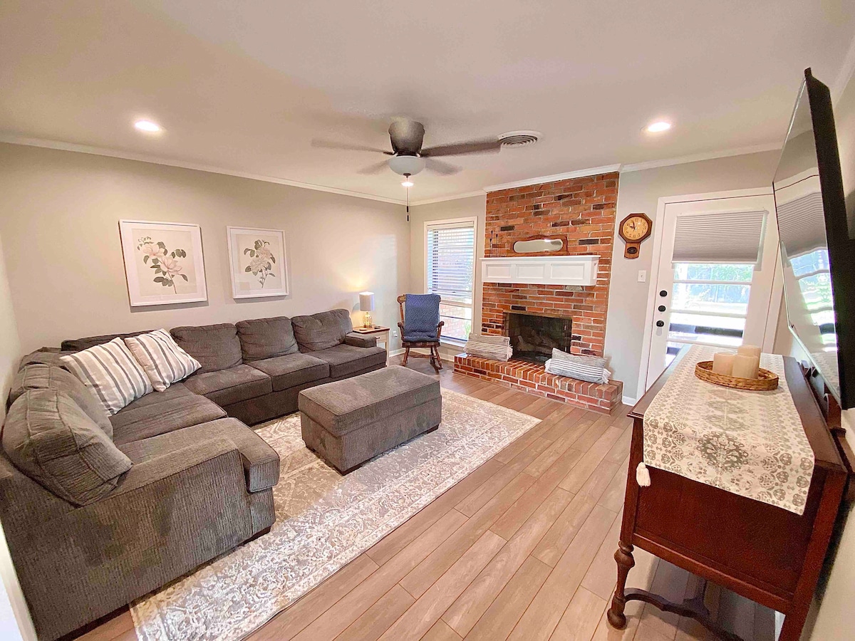 A spacious living room is highlighted by a large sectional sofa and a cozy ottoman. A brick fireplace serves as a focal point, while natural light flows in through the windows. Decorative wall art and a vintage clock add character to the inviting space.