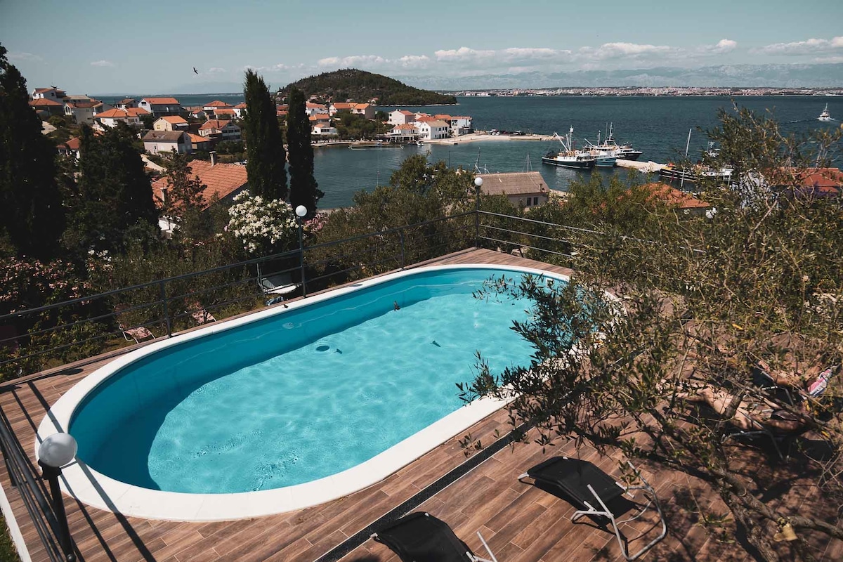 A modern swimming pool is positioned on a terrace, offering sweeping views of the coastline and the sea. Surrounding greenery and a wooden deck accommodate lounge chairs, creating a tranquil setting for relaxation. The distant shoreline features quaint houses and boats, enhancing the serene atmosphere.