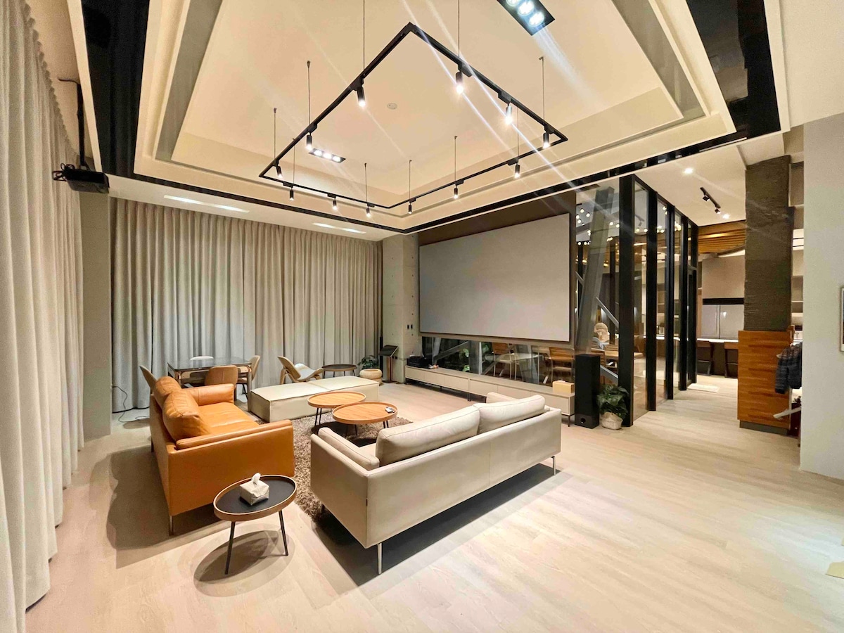 A spacious entertainment area features high ceilings and modern furnishings. A large projection screen is positioned against the wall, surrounded by comfortable seating arrangements. Soft lighting and neutral-toned curtains add to the ambient atmosphere, enhancing the inviting yet stylish space.