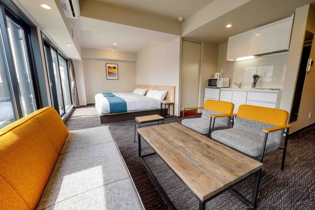 The room features a spacious layout with two large beds and a comfortable sofa in warm tones. A wooden table is centered in front of the seating area, with a kitchenette visible in the background, equipped with a microwave and other essentials.