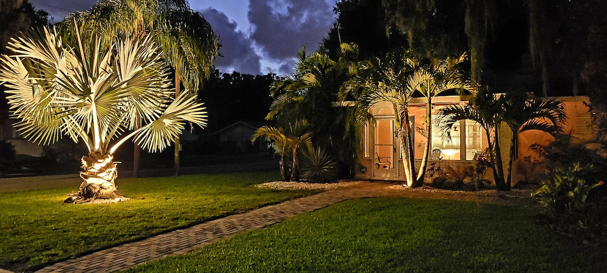 The front yard features lush greenery, highlighted by a large palm tree illuminated at night. A pathway made of pavers leads to the home, which is surrounded by additional tropical plants. Soft light illuminates the house’s exterior, creating a welcoming evening atmosphere.