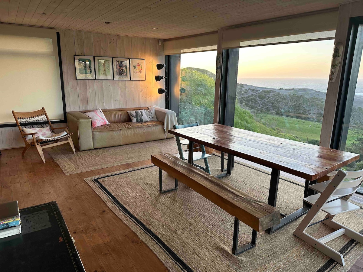 A spacious living-dining area features large glass windows offering expansive views of the surrounding landscape. A wooden table with benches is complemented by a comfortable sofa and an armchair, creating a welcoming space. Natural light fills the room, enhancing the wooden accents and neutral tones.