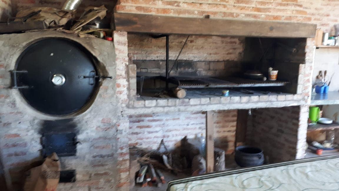 A rustic brick structure houses a wood-fired oven and a barbeque grill. Cooking utensils and a few cooking items are visible on the grill, while firewood is neatly stored beside the brickwork. The setting exudes a functional outdoor cooking space.