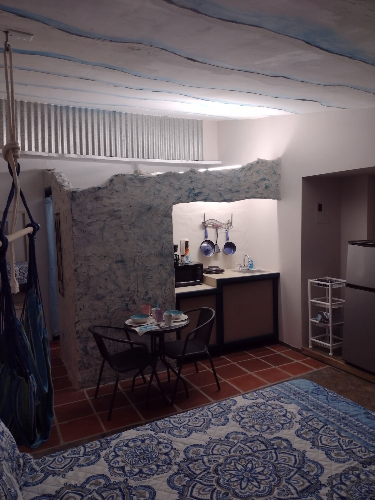 A compact living space features a kitchenette with hanging pots. A round dining table with two chairs is situated beside it, accompanied by a soft blue patterned rug. The ceiling exhibits a unique wavy design, while the walls showcase a textured finish in light blue tones.