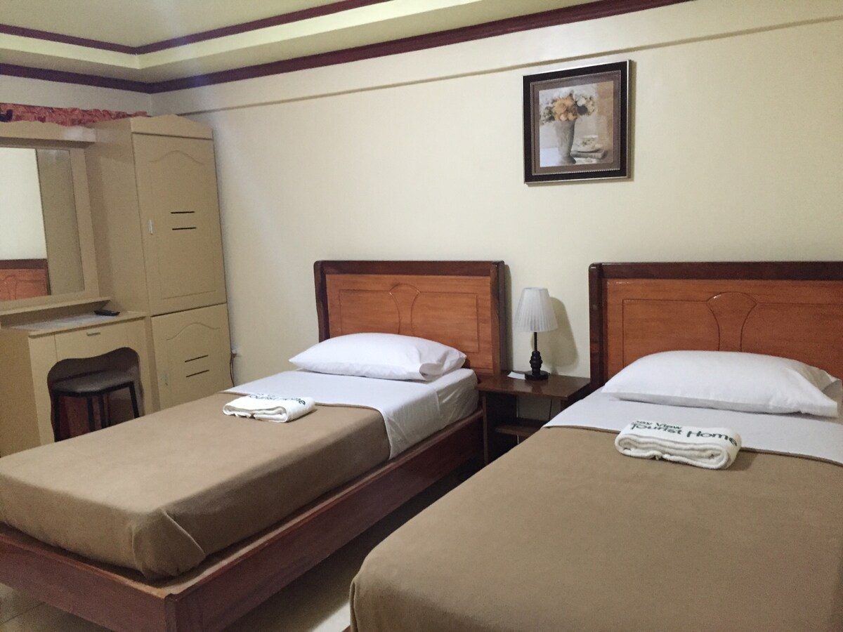 The room contains two single beds, each dressed with neutral bedding. A bedside table with a lamp is positioned between the beds. A wooden dresser and mirror are visible, along with a framed piece of wall art, adding a touch of decor.