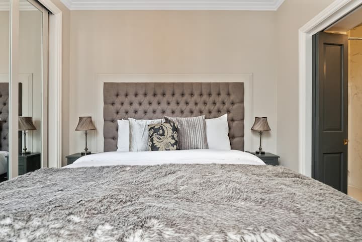 غرفة النوم الرئيسية التي توضح سريرًا بمقاس كبير.  تخزين الحائط ومساحة معلقة مخفية بجدار المرآة.