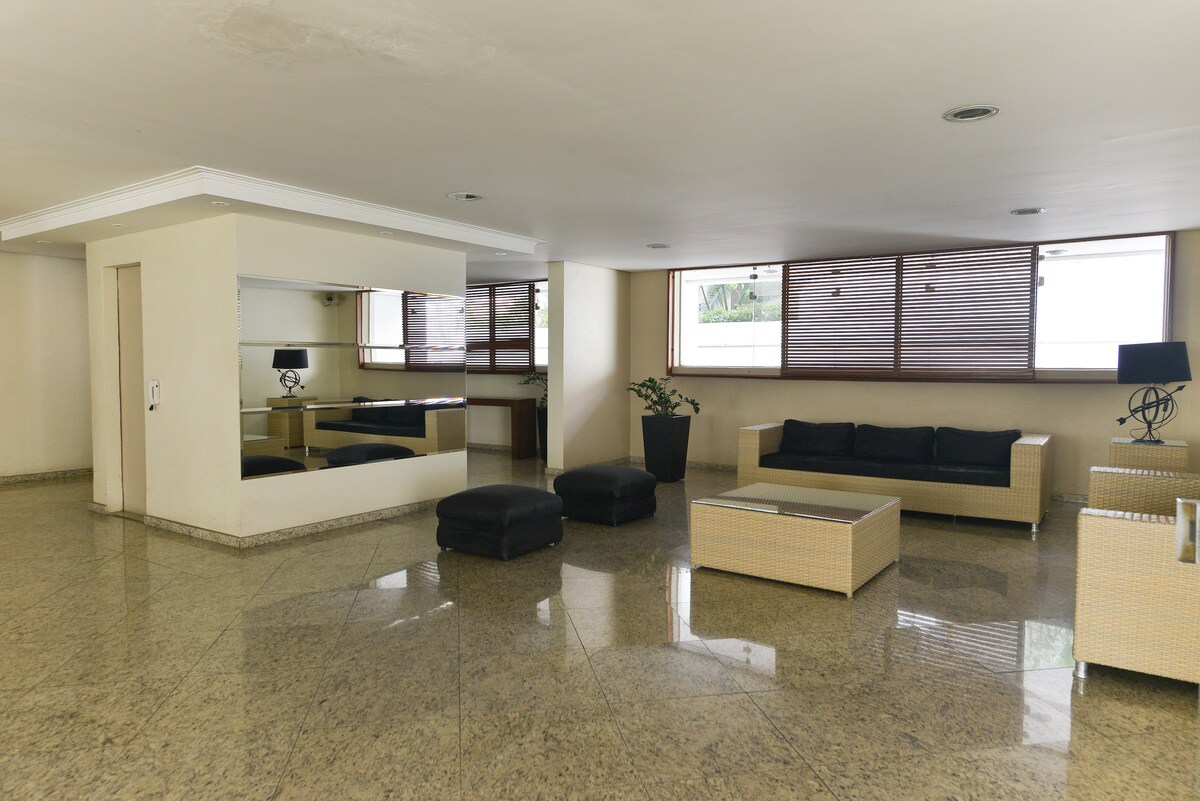 A spacious lounge area features light-colored seating arranged around a low table. Large windows allow natural light to fill the room, complemented by decorative plants and modern accent pieces. The polished flooring enhances the open and bright atmosphere of the space.