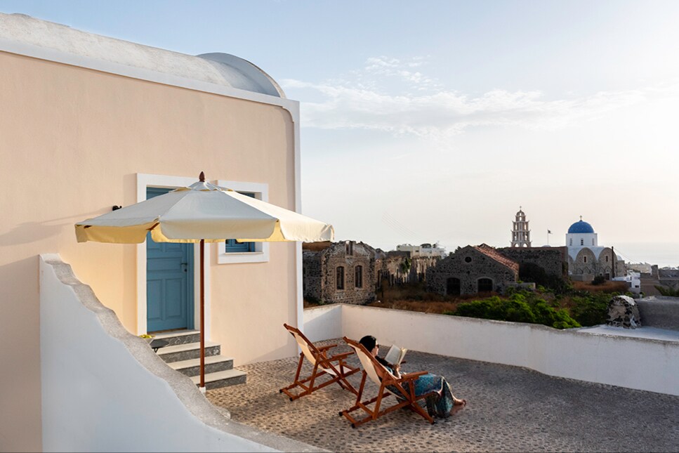 A private terrace features two lounge chairs positioned under a large umbrella, inviting relaxation. A view of historical architecture and a distant church dome is captured in the background, with soft natural light illuminating the space.