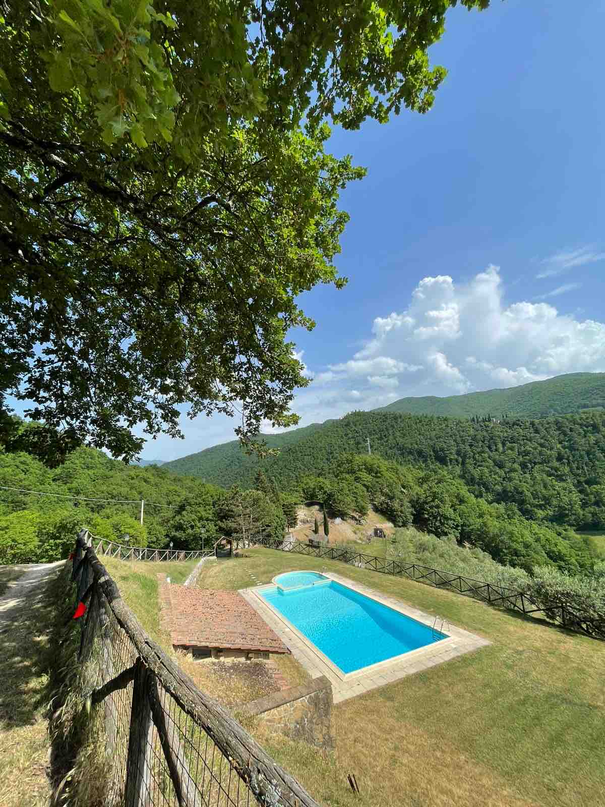 A serene outdoor scene showcases a swimming pool surrounded by lush greenery and rolling hills. The pool is framed by a wooden fence and a well-kept lawn, under a clear blue sky dotted with fluffy clouds, inviting relaxation in a tranquil environment.