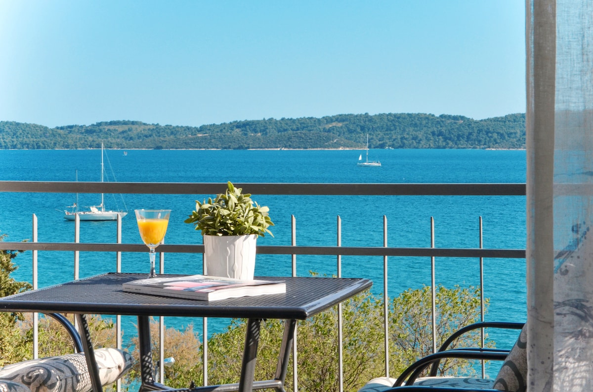 A balcony overlooks a serene blue sea, featuring a small table with a glass of orange juice and a potted plant. Sailboats can be seen in the distance against a clear sky, creating a peaceful coastal scene.