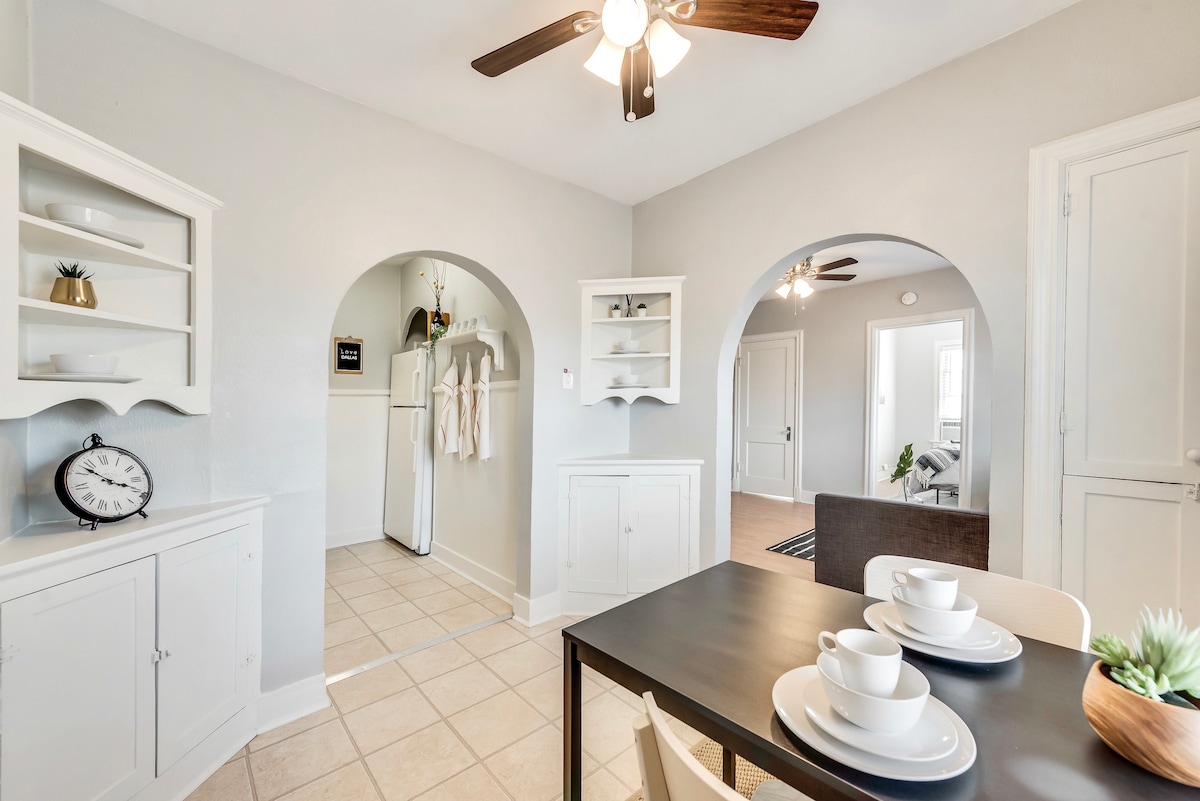 The dining area features a simple table set for two, with white dishes and cups. Light-colored cabinets outline the space, and arched doorways lead to the kitchen and living area. A ceiling fan provides a functional element amidst the neutral wall tones.
