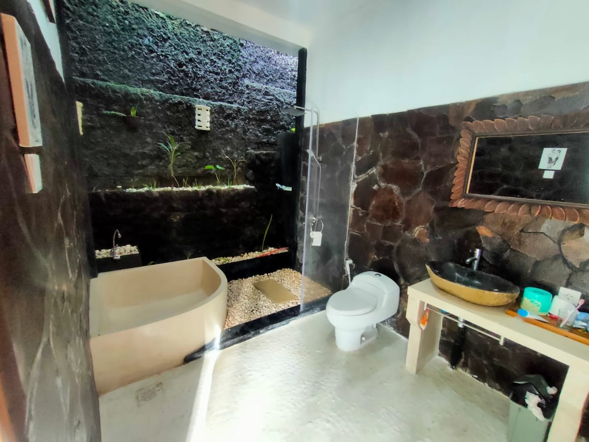 A modern bathroom features a combination of natural stone walls and a sleek design. A spacious bathtub is situated beside a shower area, both surrounded by an indoor garden with plants. A wooden sink area is equipped with a round basin and complementary toiletries.