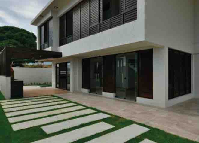 The exterior of the house is shown, featuring a modern design with large glass doors and sleek wooden shutters. A smooth stone pathway leads to the entrance, bordered by a well-maintained grassy area, creating an open and inviting entryway.
