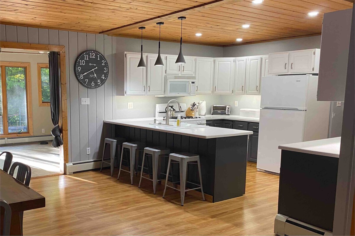 A spacious kitchen is designed with modern amenities and a large island featuring seating for four. Light wood ceilings contrast with gray cabinetry, while natural light floods in through large windows. Essentials like a microwave and refrigerator are included for convenience.