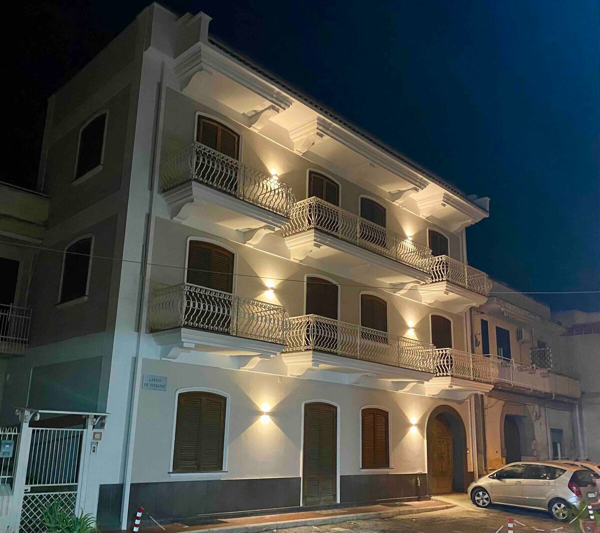 A three-story building is illuminated at night, highlighting its architectural details. Balconies with decorative railings can be seen along each floor. The entrance is framed by elegant columns, and nearby cars are parked along the street, enhancing the building's inviting appearance.