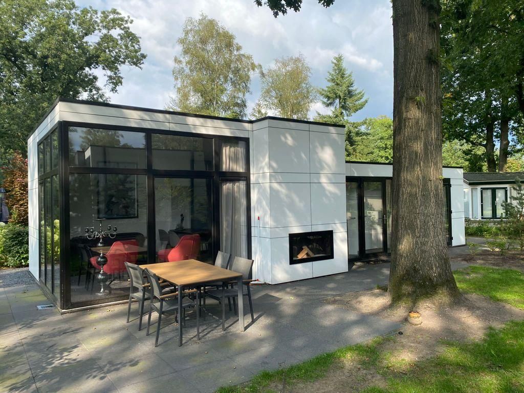 A modern single-story structure is surrounded by lush greenery, featuring large glass panels that allow natural light to flood the interior. An outdoor dining area with a table and chairs is positioned on a stone patio, providing a relaxed space for meals.