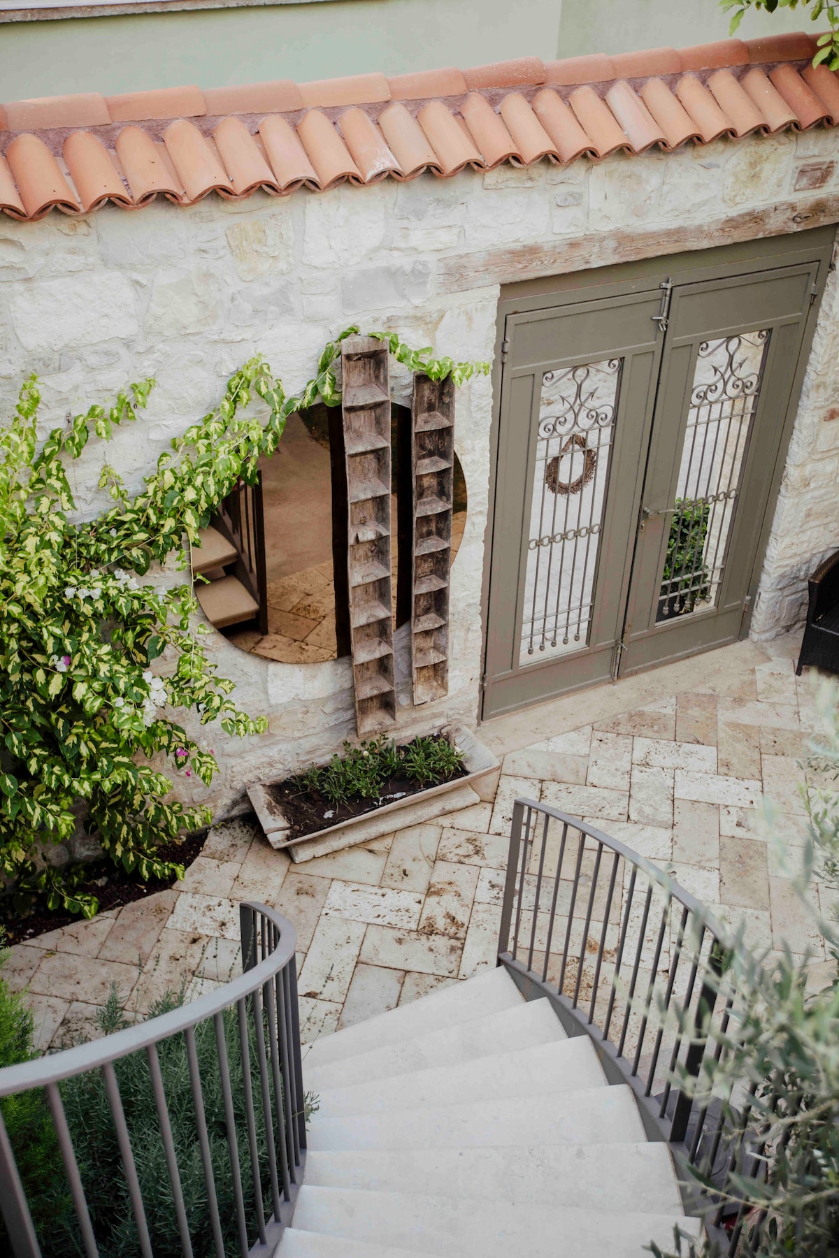 A charming outdoor area is framed by a stone wall and features a staircase leading down. Green foliage climbs up the side of a wall, while a decorative wooden shelf adds character. An arched window and double doors provide access to the interior.