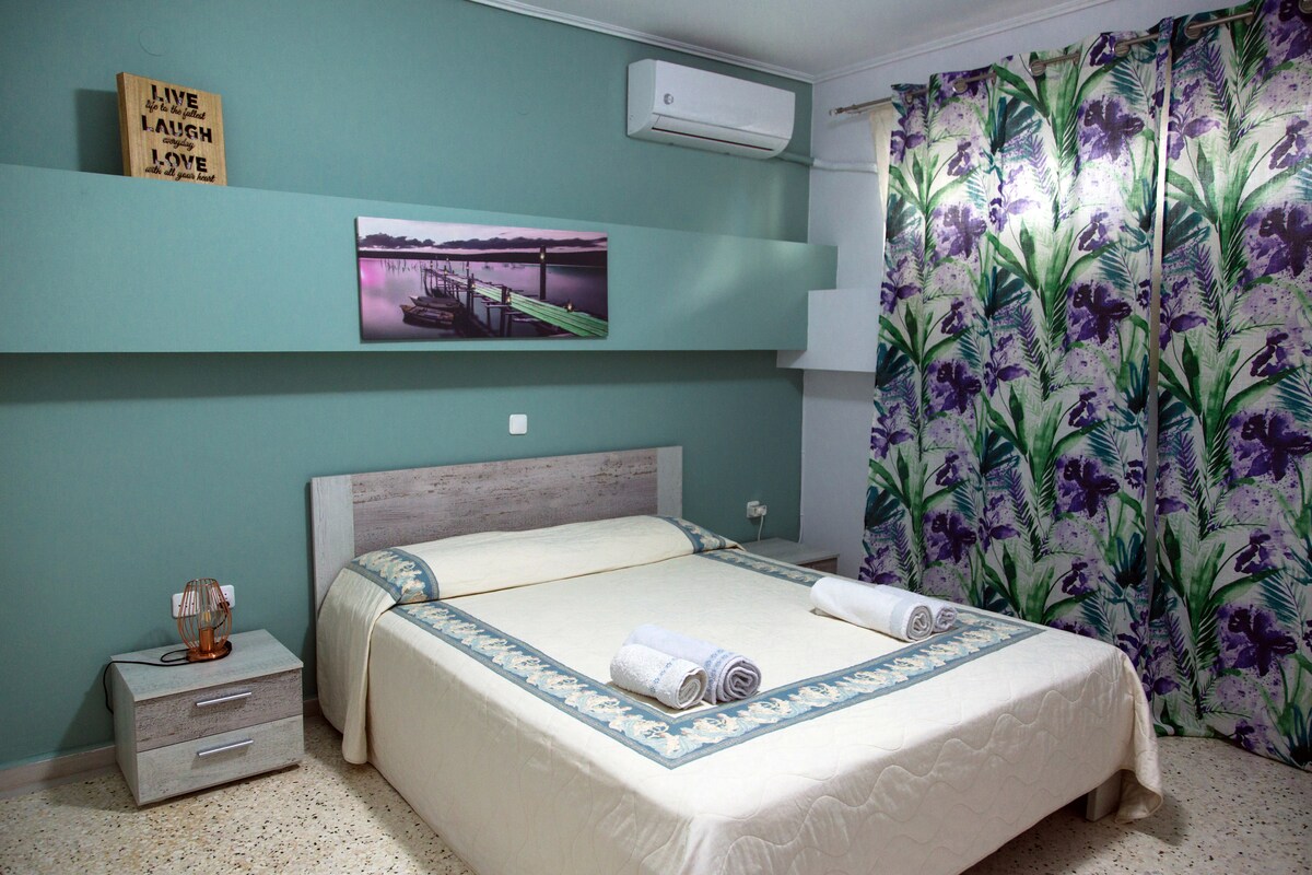 A spacious sleeping area is presented with soft, neutral-toned bedding on a large bed. Decorative towels are neatly folded at the foot of the bed. A wall-mounted air conditioning unit provides climate control, while vibrant curtains with floral designs add a touch of color to the room.