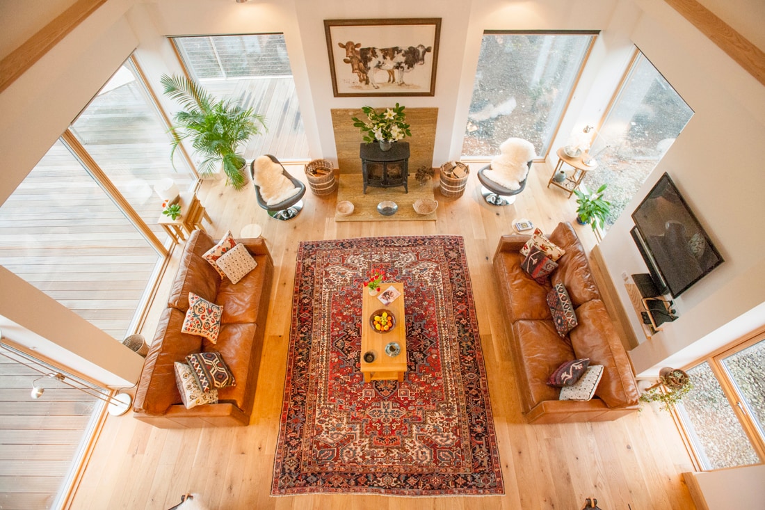 An open-concept living area features two seating arrangements on either side of a central wooden coffee table. A vibrant area rug adds warmth to the space, while large windows flood the room with natural light, enhancing the views of the surrounding environment.
