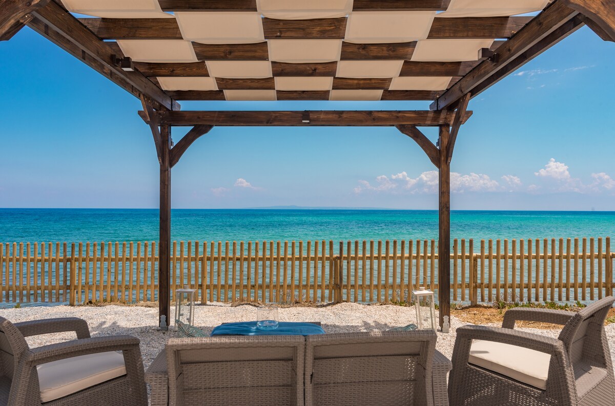 A shaded outdoor seating area features comfortable chairs arranged in front of a stunning view of the blue-green waters and sandy beach. A wooden pergola overhead provides sun protection, while a wooden fence outlines the beachfront location.