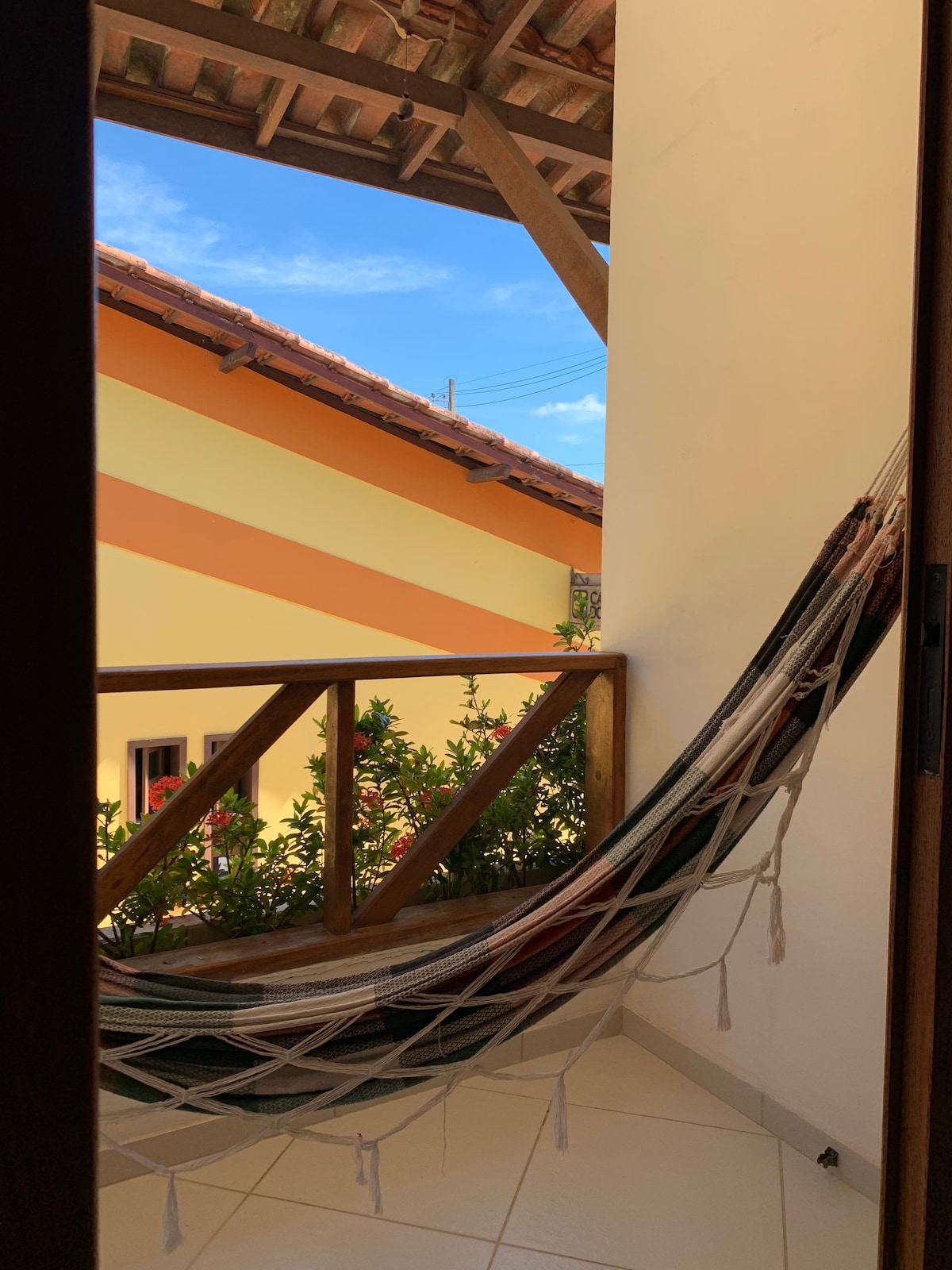 A private balcony features a cozy hammock hanging between wooden posts. Lush green plants are visible below, along with a view of neighboring buildings with warm-colored façades. The sky is clear, adding a sense of openness and calmness to the outdoor space.