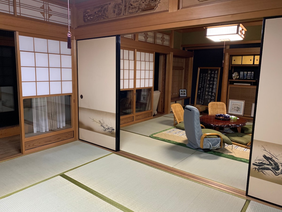 A traditional Japanese room is featured, with sliding shoji doors leading to a tatami-matted space. A low wooden table is surrounded by wicker chairs, while decorative wall art enhances the ambiance. Natural light filters through the windows, highlighting the room's simplicity and elegance.