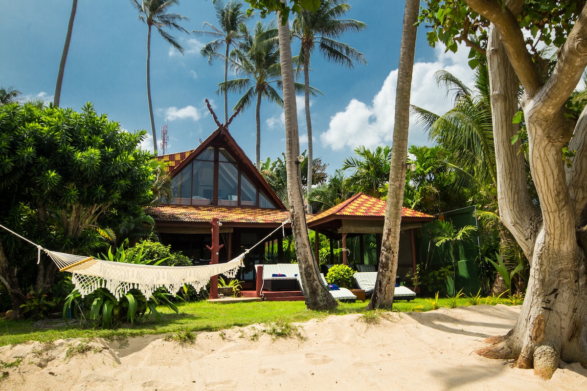 A Thai-style wooden villa is framed by tropical foliage and palm trees. A sandy area leads to a inviting hammock swaying gently between two trees. The unique rooftop structure showcases wooden details, with loungers positioned for relaxation under the sun.