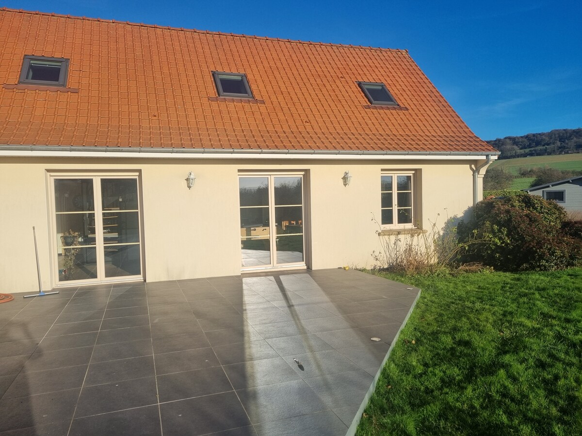 The exterior of the house showcases a light-colored façade complemented by a red tiled roof. Large glass doors provide access to a paved terrace. Surrounding greenery and a well-maintained lawn add to the tranquil atmosphere, while mountains can be seen in the background.