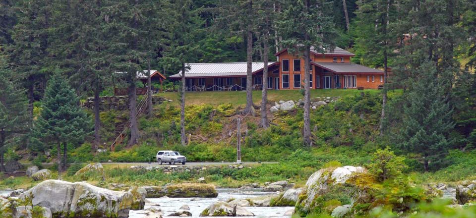 A spacious lodge is set amidst tall trees beside the Chilkoot River. The building features large windows and a multi-level design. A vehicle is parked along the nearby gravel road, with natural rock formations and greenery surrounding the property.