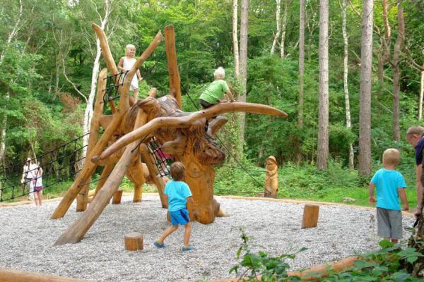 A wooden playground structure resembling a large animal is surrounded by a gravel surface in a forested area. Children are seen climbing and playing on the structure, while others explore nearby. Lush green trees provide a natural backdrop, enhancing the outdoor play experience.