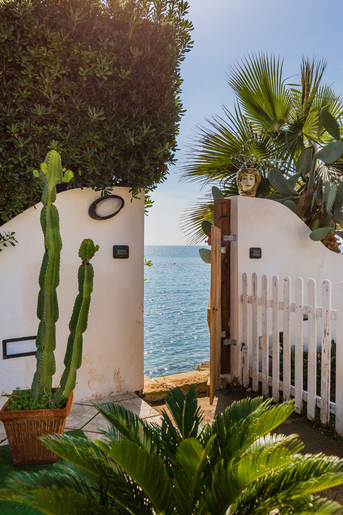 A garden gate opens to reveal a serene view of the azure sea. Lush greenery, including a tall cactus and palm trees, frames the scene, enhancing the coastal ambiance. The white gate contrasts with the natural elements, inviting guests to explore the waterfront.