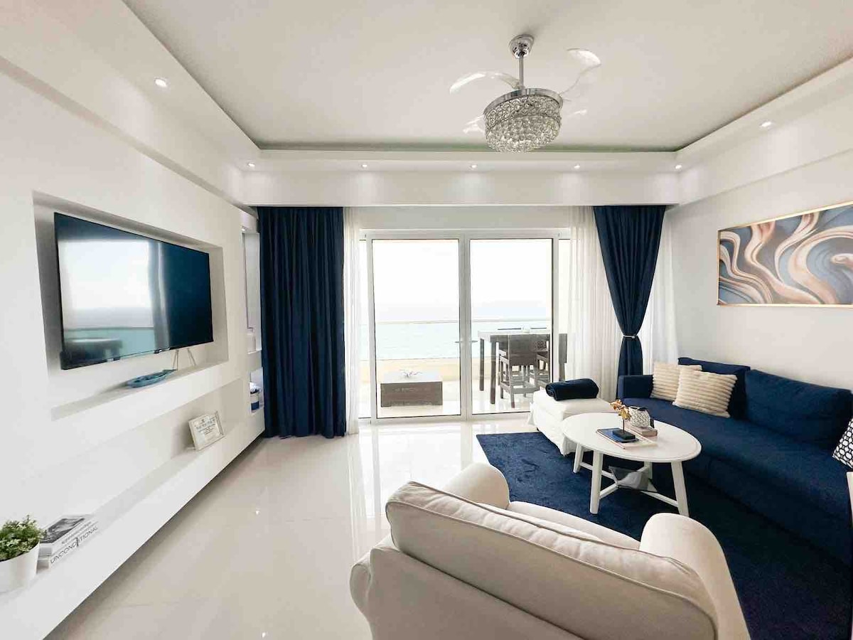 A modern living area is showcased, featuring a comfortable blue sofa and a white coffee table. A large flat-screen TV is mounted on the wall. Floor-to-ceiling glass doors provide a view of the ocean, with sheer curtains allowing natural light to fill the space.