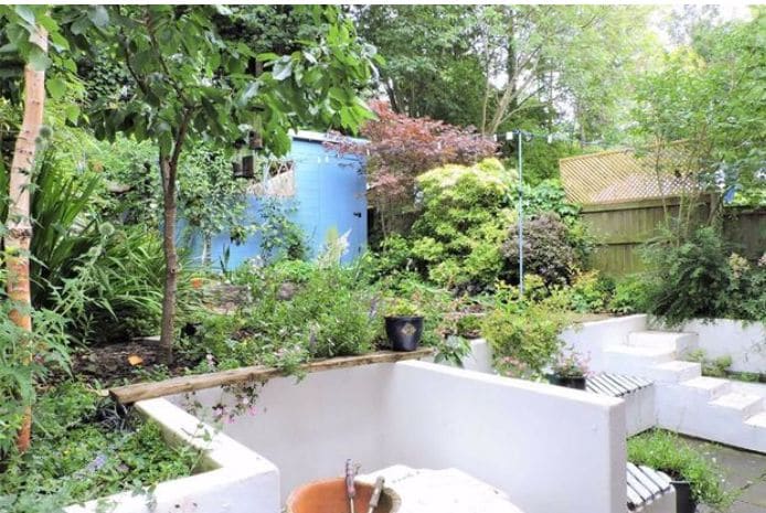 A serene garden space is surrounded by lush greenery, showcasing various plants and trees. A quaint blue shed is visible in the background, while several white raised beds create distinct planting areas. A peaceful atmosphere is enhanced by the natural beauty of the setting.