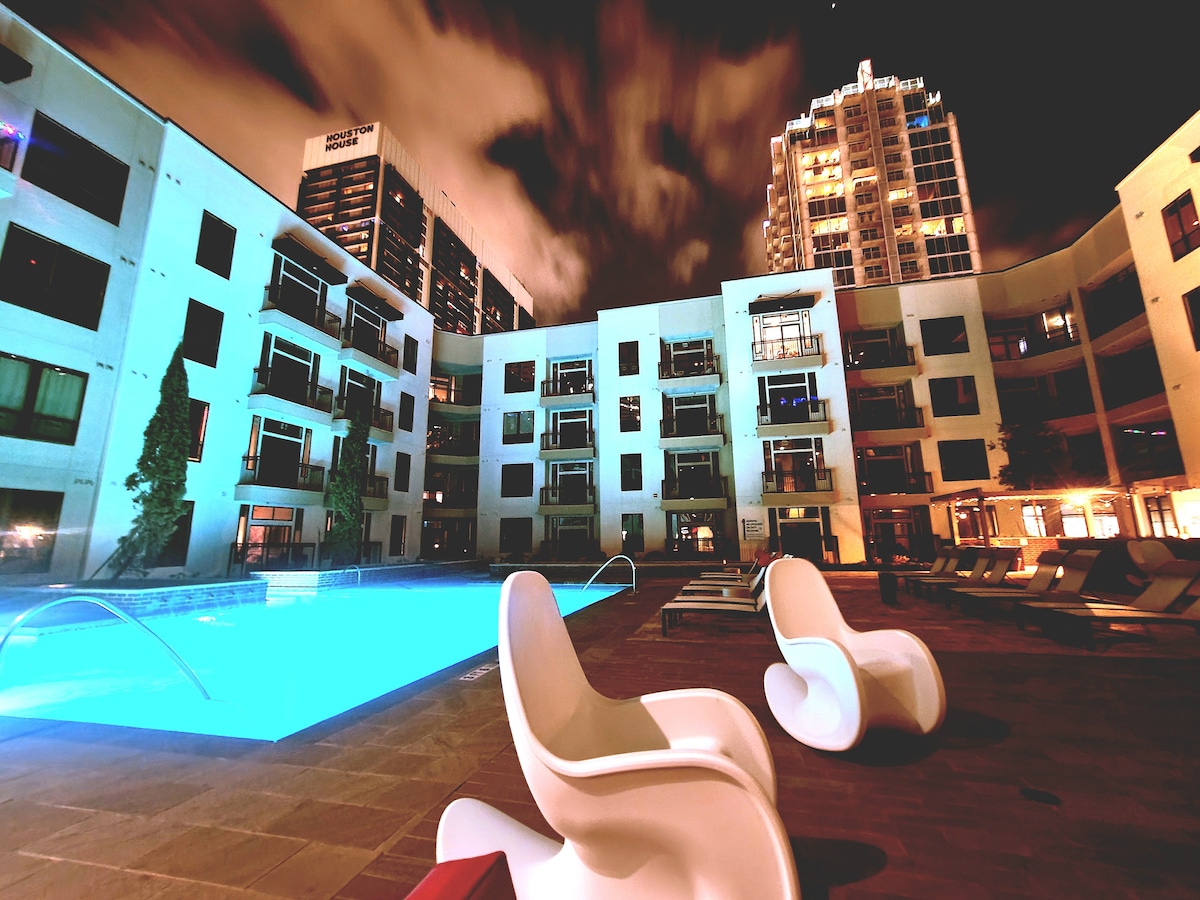 An outdoor pool area is illuminated at night, showcasing sleek white lounge chairs positioned beside the tranquil water. Surrounding buildings reflect soft lighting, creating a serene atmosphere. The pool is accentuated by a light-colored stone deck and overlooks an inviting space for relaxation.