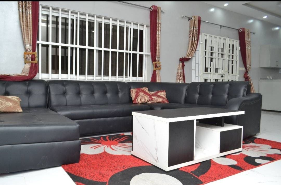 A spacious living room features a large black sectional sofa adorned with decorative cushions. A coffee table with a marble finish sits on a vibrant area rug, while large windows allow natural light to fill the space, complemented by stylish curtains.