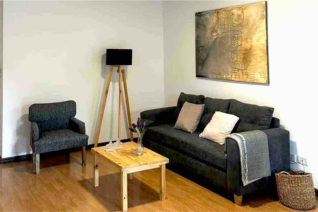 A contemporary living area features a dark gray sofa with light decorative pillows, accompanied by a patterned armchair. A wooden coffee table holds fresh flowers, and a floor lamp adds subtle illumination. A large wall art piece creates visual interest above the sofa.