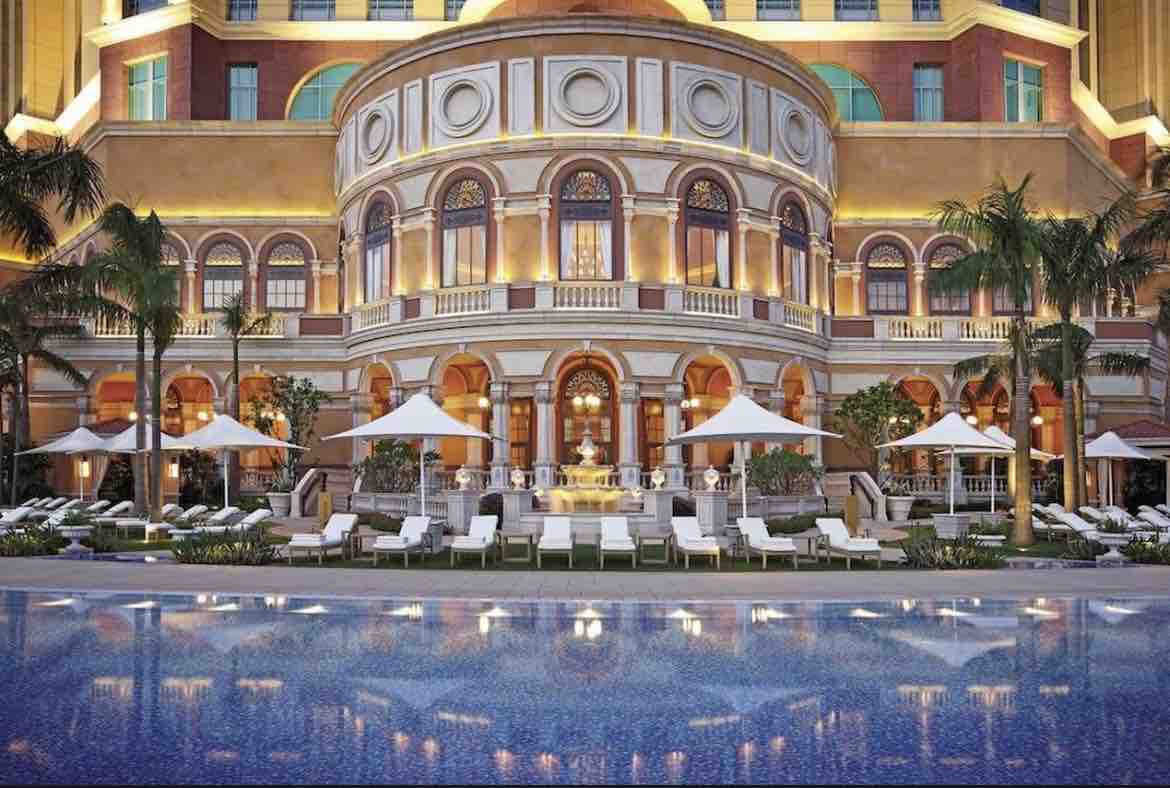 The elegant exterior of a high-end property is showcased, featuring a grand façade with arched windows and decorative detailing. A pool reflects the building's architecture, surrounded by lounge chairs and umbrellas. Lush greenery and well-kept landscaping enhance the overall setting.