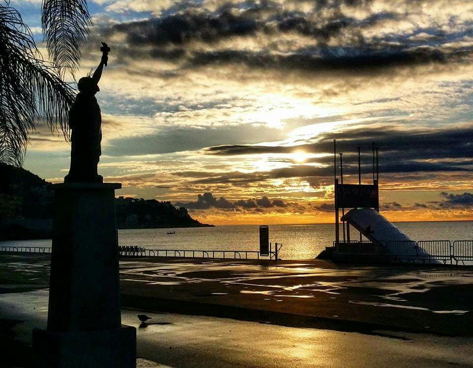 A silhouette of a statue reminiscent of the Statue of Liberty stands by the waterfront, outlined against a cloudy sunset. The sea glimmers in the fading light, while the shoreline is lined with a low barrier and visible structures in the distance.