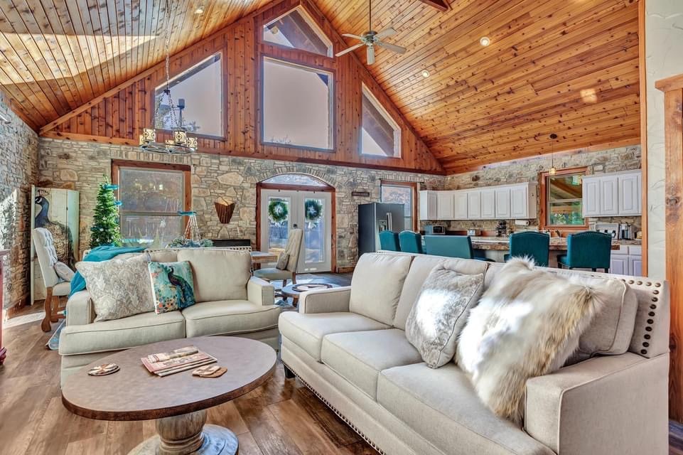 A spacious living area features vaulted wood ceilings and large windows, allowing natural light to flow in. Comfortable seating is arranged around a round coffee table, with decorative elements thoughtfully placed throughout. The open layout extends to a dining area and kitchen visible in the background.