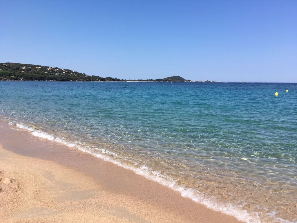 A sandy beach is depicted, featuring gentle waves lapping against the shore. The clear blue water displays varying shades, with a view of distant hills and lush greenery along the coastline. A bright sky without clouds enhances the serene coastal scene.