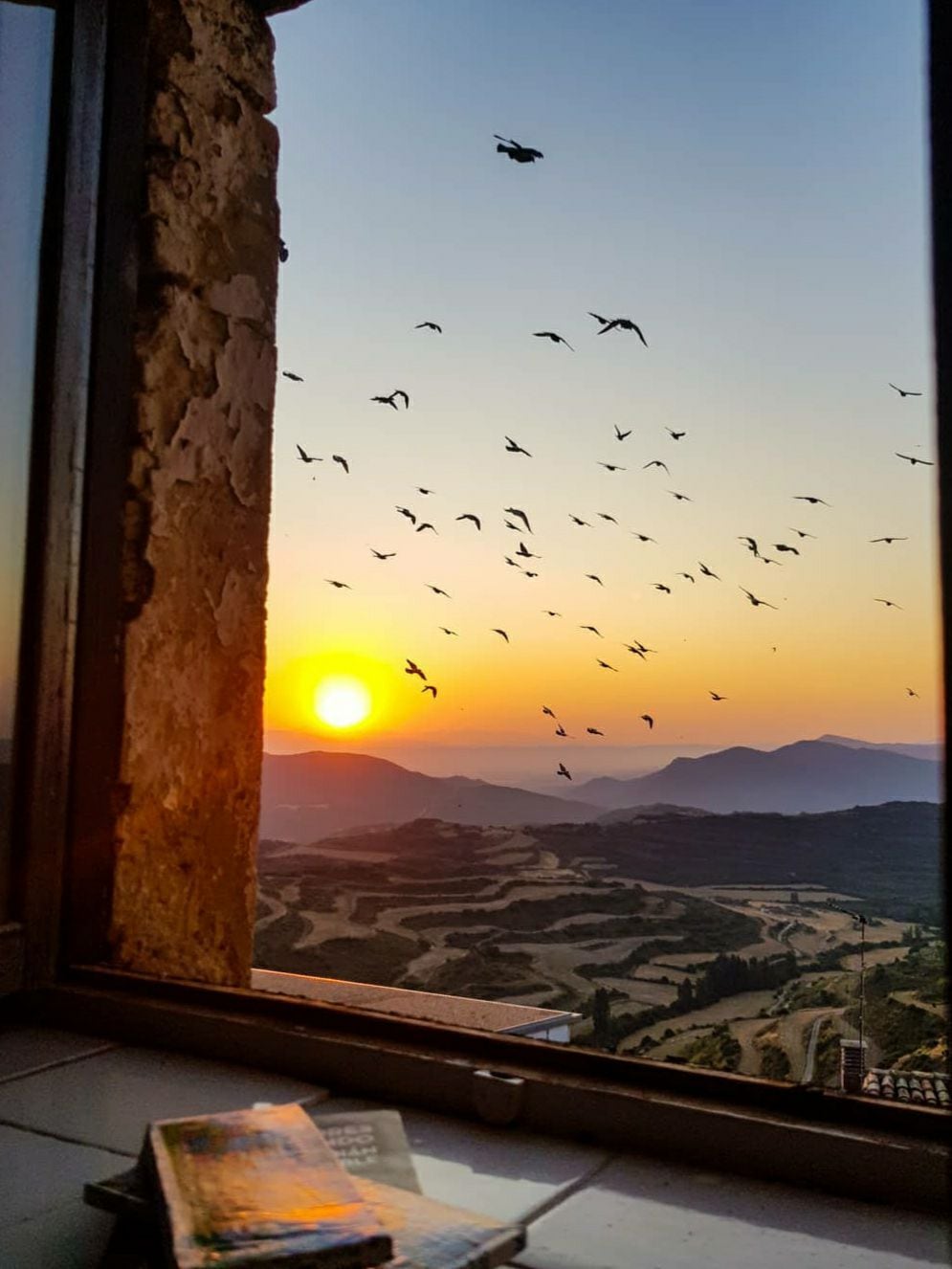 A window frames a stunning sunset with the sun dipping below rolling hills. Silhouetted birds can be seen flying through the warm orange and yellow sky, while a few books rest on a surface below, hinting at quiet moments inside.