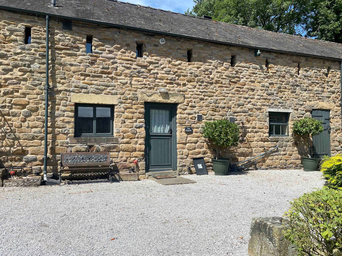 The exterior of the barn conversion features a rustic stone façade, punctuated by small windows and a green door. A gravel pathway leads to the entrance, complemented by potted plants on either side. A wooden bench is situated nearby, inviting relaxation.