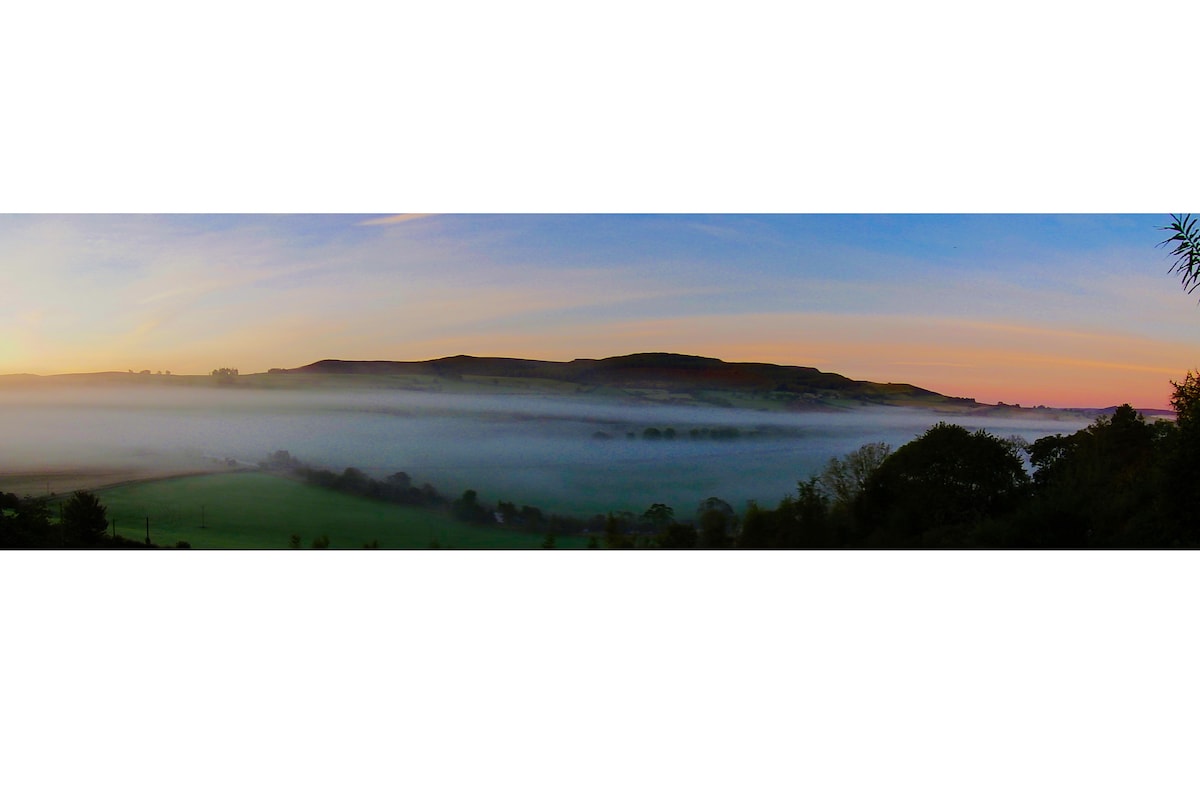 A panoramic view captures a misty landscape at dawn, with soft hues of pink and orange illuminating the sky. The rolling hills are partially shrouded in fog, while the lush green fields below create a serene and tranquil atmosphere.