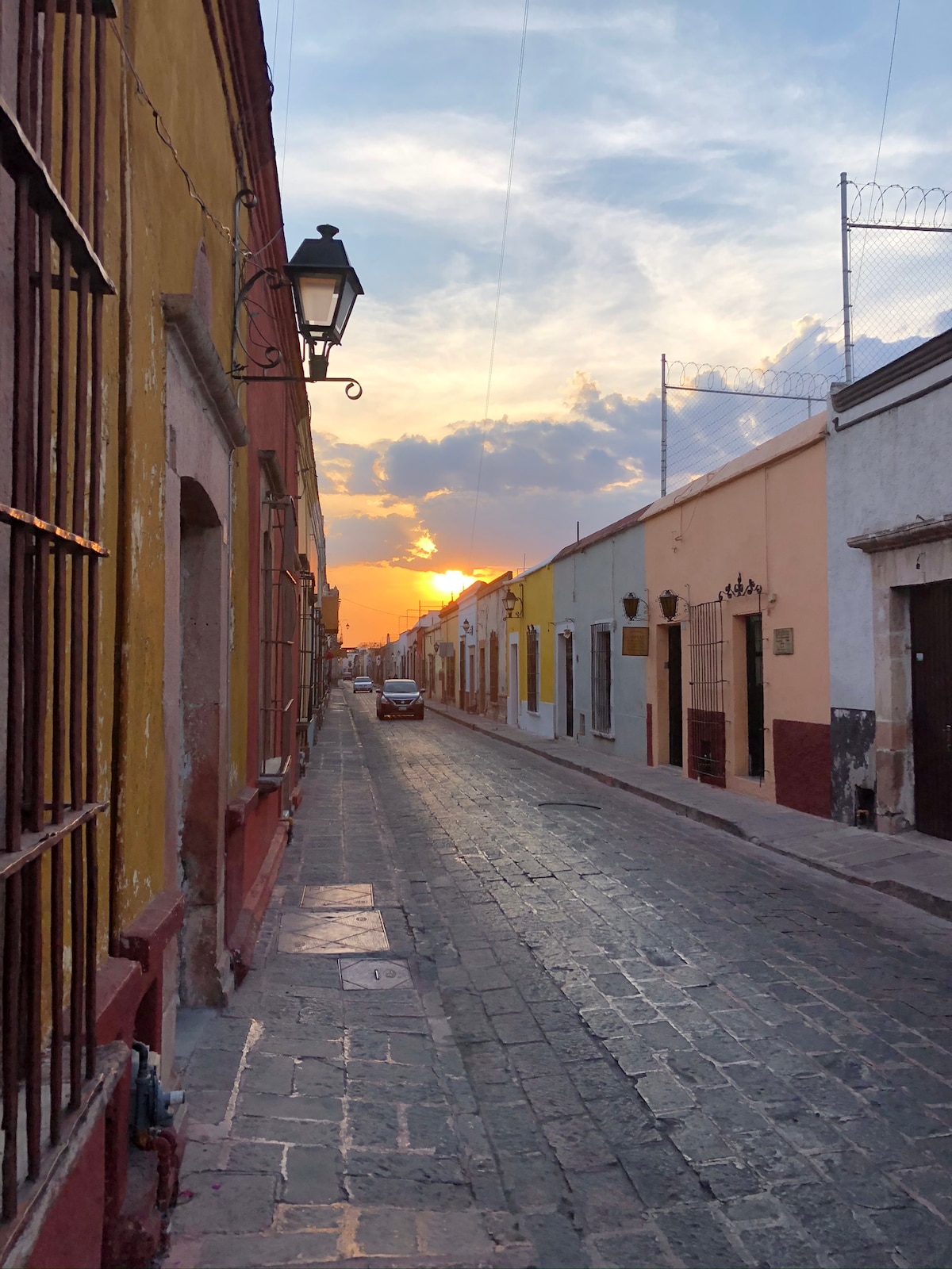 A quiet street in Querétaro is captured at sunset, with warm tones illuminating the colorful facades of historic buildings on either side. Cobblestone pavement leads toward the horizon where the sun sets, casting a gentle glow over the tranquil scene.