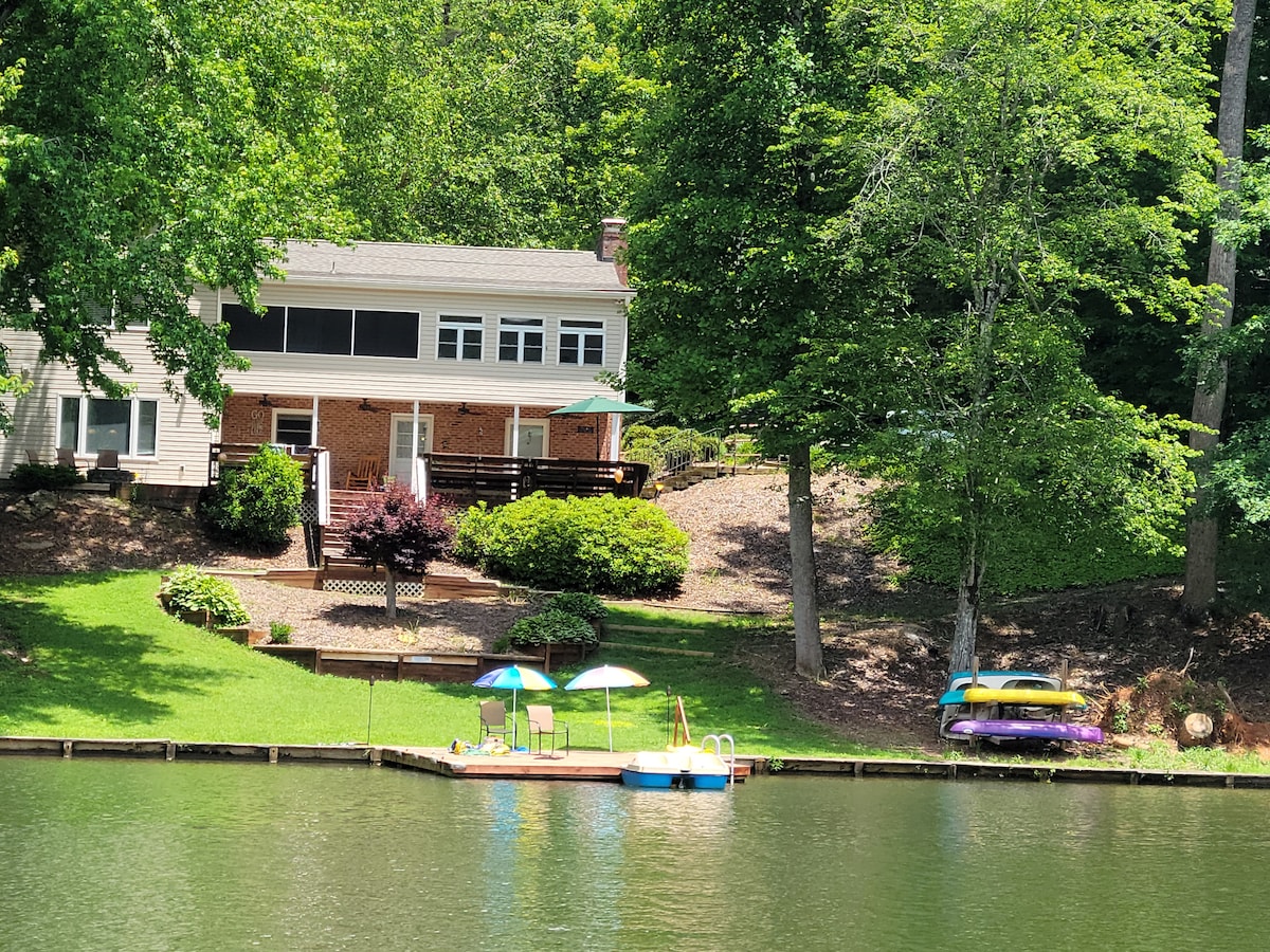 The image showcases a lakefront home surrounded by greenery, with a spacious lawn leading down to the dock. A floating dock features umbrellas and seating, while colorful kayaks rest alongside. The home, with multiple levels, presents a welcoming, nature-filled setting.