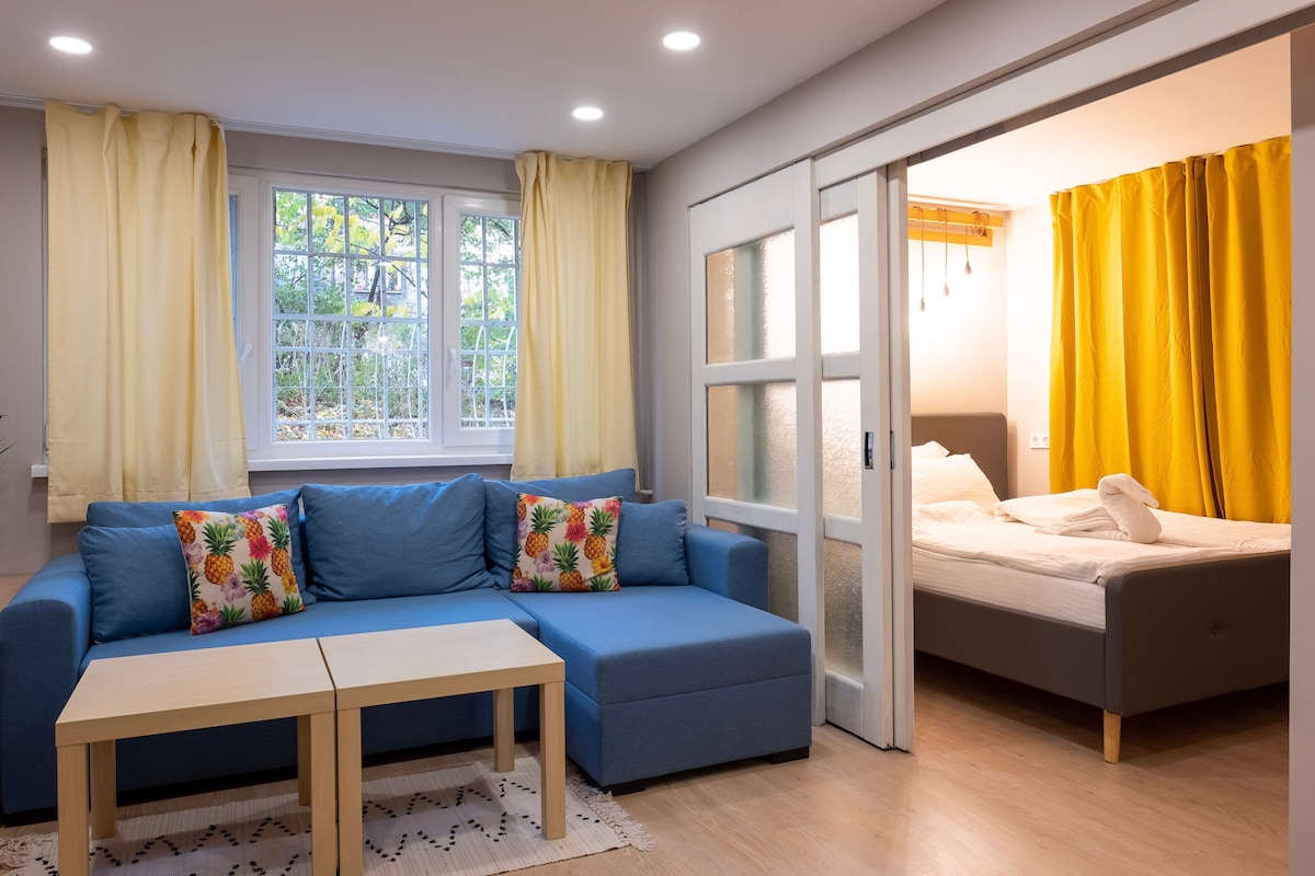 The living area features a blue sofa adorned with colorful pineapple-patterned cushions, accompanied by a wooden coffee table. A doorway leads to a well-lit bedroom with warm yellow curtains, revealing a neatly made bed in the background.
